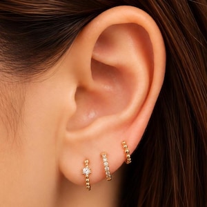 May include: Three gold hoop earrings worn in a row on an earlobe. The first hoop features a single clear stone with beaded detailing, the second is a pavé-style hoop with multiple small clear stones, and the third is a simple gold beaded hoop.