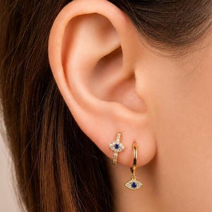 May include: A close-up of a person's ear adorned with two gold earrings. One is a small hoop with a dangling blue eye charm. The other is a small hoop with a blue eye design. Both earrings are embellished with small, clear stones.