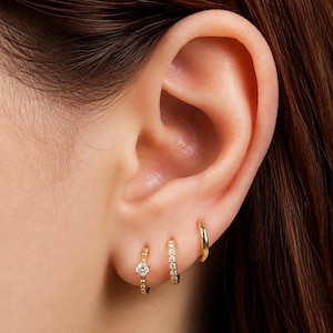 May include: Three gold hoop earrings worn in a single earlobe. The first hoop features a single clear stone, the second hoop is lined with multiple small clear stones, and the third hoop is a plain, smooth gold band without any stones.