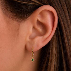 Peut inclure: Une boucle d'oreille créole en or avec une petite breloque pendante composée d'un cadre carré et d'une minuscule pierre précieuse ronde d'un vert éclatant au centre. La boucle est fixée au lobe par un anneau fin en métal doré poli.