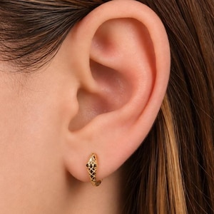 May include: A gold hoop earring shaped like a snake, featuring a textured, lattice-like pattern across its body and two small, dark, circular stones set as eyes. The earring is worn in the lower earlobe, showcasing its curved, slender design.