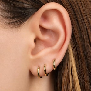 May include: Three small, polished gold hoop earrings are worn in a row along the earlobe. Each earring features a simple, circular design with a smooth, reflective metallic finish. The hoops are graduated in size, with the largest positioned highest.