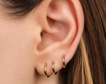 May include: Three small, polished gold hoop earrings are worn in a row along the earlobe. Each hoop features a smooth, circular design with a reflective metallic finish. The earrings are graduated in size, with the smallest hoop positioned at the bottom and the largest hoop placed highest on the earlobe.