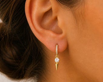 May include: Gold-toned hoop earring with a small, clear gemstone and a spike pendant. The hoop is encrusted with small, clear gemstones. The earring is shown on a person's ear.