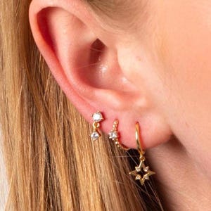 May include: A close-up of a person's ear adorned with three sets of gold earrings. The earrings include small studs with dangling crystals, a chain of small beads, and a hoop earring with a star pendant.