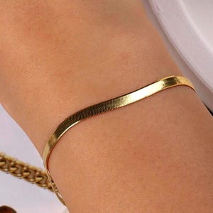 May include: A gold-colored, flat herringbone bracelet is worn on a wrist. The bracelet has a smooth, reflective surface. A gold chain is visible in the background. The bracelet is a piece of jewelry.