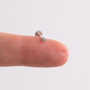 May include: A small, silver-colored stud earring with a clear, round gemstone. The earring has a flat back and a post. The gemstone is set in a bezel setting. The earring is shown on a fingertip against a white background.