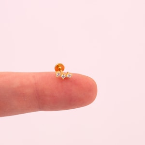 May include: A small, gold-coloured earring featuring three clear gemstones. The earring is designed for a lip or cartilage piercing. The post is a standard size for body jewellery.