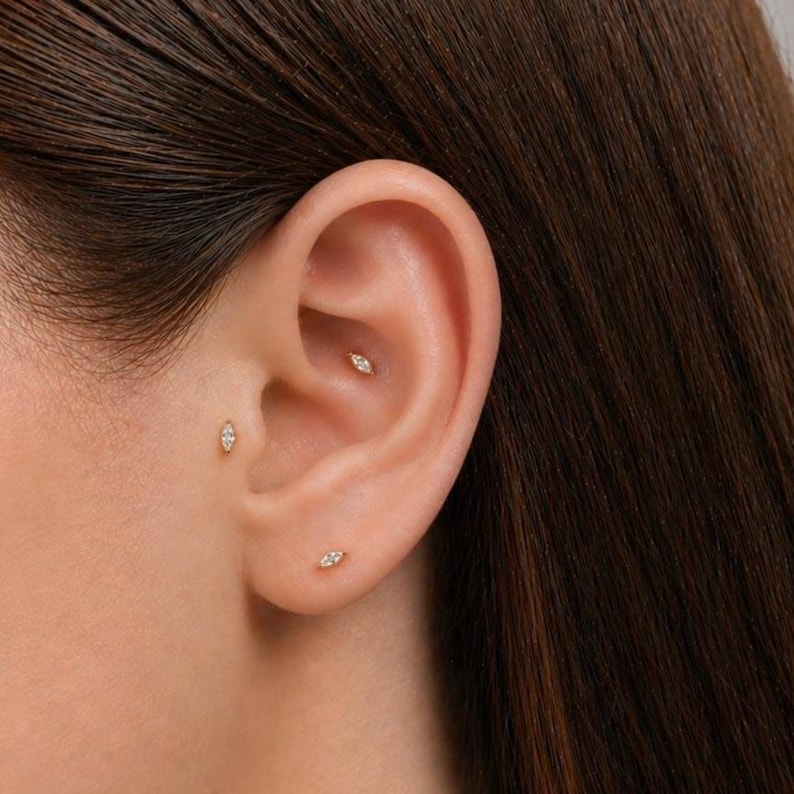 May include: Three small gold stud earrings, each featuring a marquise-cut clear gemstone. One earring is placed in the earlobe, one in the tragus, and one in the conch area. The earrings have a delicate, elongated shape and a polished metallic finish.