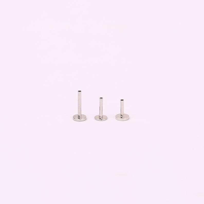 May include: Three silver-toned metal labret studs arranged in a row, each featuring a flat circular base and a hollow cylindrical post of varying heights, designed for body piercing jewelry with a smooth, polished, and reflective metallic finish.