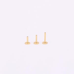May include: Three gold-coloured labret studs with flat backs, in varying lengths. Each stud features a circular base and a straight post. The studs are arranged from shortest to longest, showcasing a minimalist design. The background is plain white.