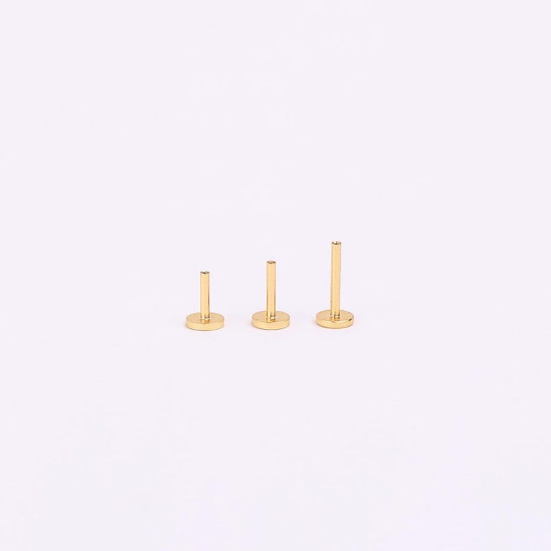 May include: Three gold-coloured labret studs with flat backs, in varying lengths. Each stud features a circular base and a straight post. The studs are arranged from shortest to longest, showcasing a minimalist design. The background is plain white.