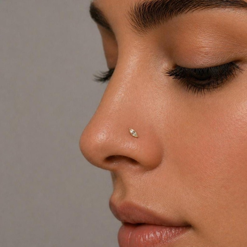 May include: A small, marquise-shaped nose stud featuring a clear, faceted gemstone set in a metallic frame. The jewelry is positioned on the side of the nostril, showcasing a slender, elongated design that sits flush against the skin surface.