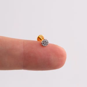 May include: A small, gold-colored stud earring with a cluster of clear, sparkling stones. The earring is round and sits on a flat post. The jewelry is shown against a neutral background.