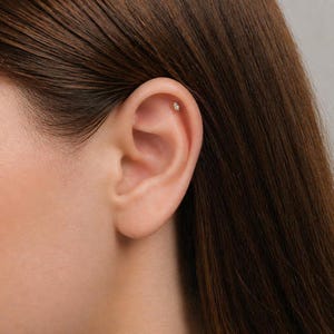 May include: A small, marquise-cut clear gemstone stud earring is positioned in the upper cartilage of the ear. The earring features a metallic setting that holds the elongated, pointed stone securely in place against the skin of the ear.