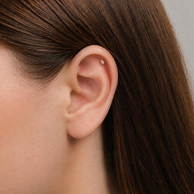 May include: A small, marquise-cut clear gemstone stud earring is positioned in the upper cartilage of the ear. The earring features a metallic setting that holds the elongated, pointed stone securely in place against the skin of the ear.