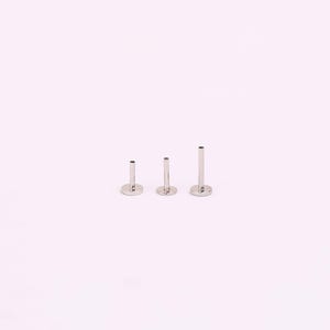 May include: Three silver-coloured flat back labret studs of varying lengths. Each stud has a flat, circular base and a straight post. The studs are arranged in a row, showing the size differences. The background is a solid, light pink colour.