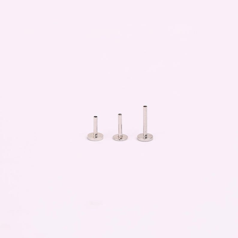 May include: Three silver-coloured flat back labret studs of varying lengths. Each stud has a flat, circular base and a straight post. The studs are arranged in a row, showing the size differences. The background is a solid, light pink colour.