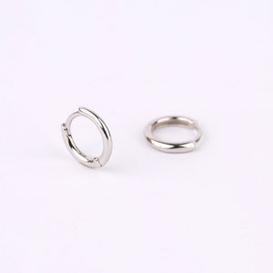 May include: A pair of small, polished silver-tone hoop earrings. One earring is positioned upright, showing the circular shape and hinged closure mechanism, while the other earring lies flat on its side, displaying the smooth, rounded metal surface.