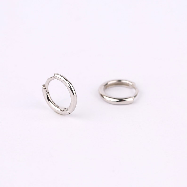 May include: A pair of small, polished silver-tone hoop earrings. One earring is positioned upright, showing the circular shape and hinged closure mechanism, while the other earring lies flat on its side, displaying the smooth, rounded metal surface.