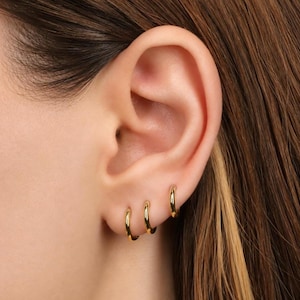 May include: Three small, polished gold hoop earrings are worn in a row along the lower edge of the earlobe. The hoops are circular, smooth, and metallic, featuring a simple, continuous design that sits flush against the skin of the ear.