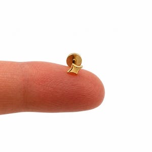 May include: A gold-coloured stud earring featuring a four-pointed star shape with a flat, circular backing. The metal surface is smooth and reflective, showcasing a polished finish on the star-shaped front and the round base of the jewellery piece.