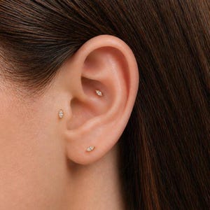 May include: Close-up of an ear with three small, gold earrings. Each earring has a small, marquise-shaped design with a central, sparkling stone. The earrings are placed in different locations on the ear.