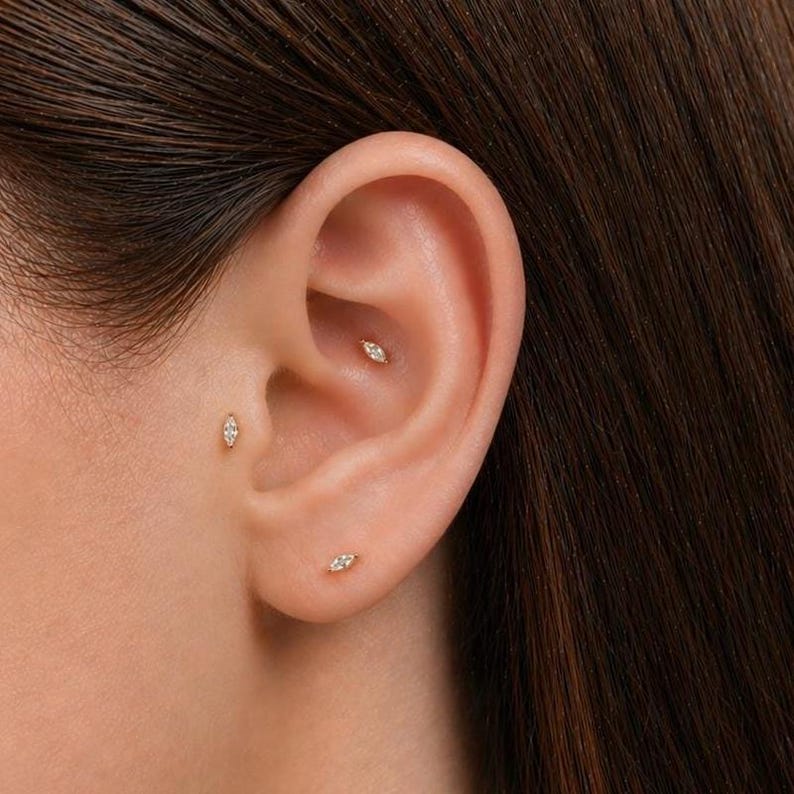 May include: Close-up of an ear with three small, gold earrings. Each earring has a small, marquise-shaped design with a central, sparkling stone. The earrings are placed in different locations on the ear.