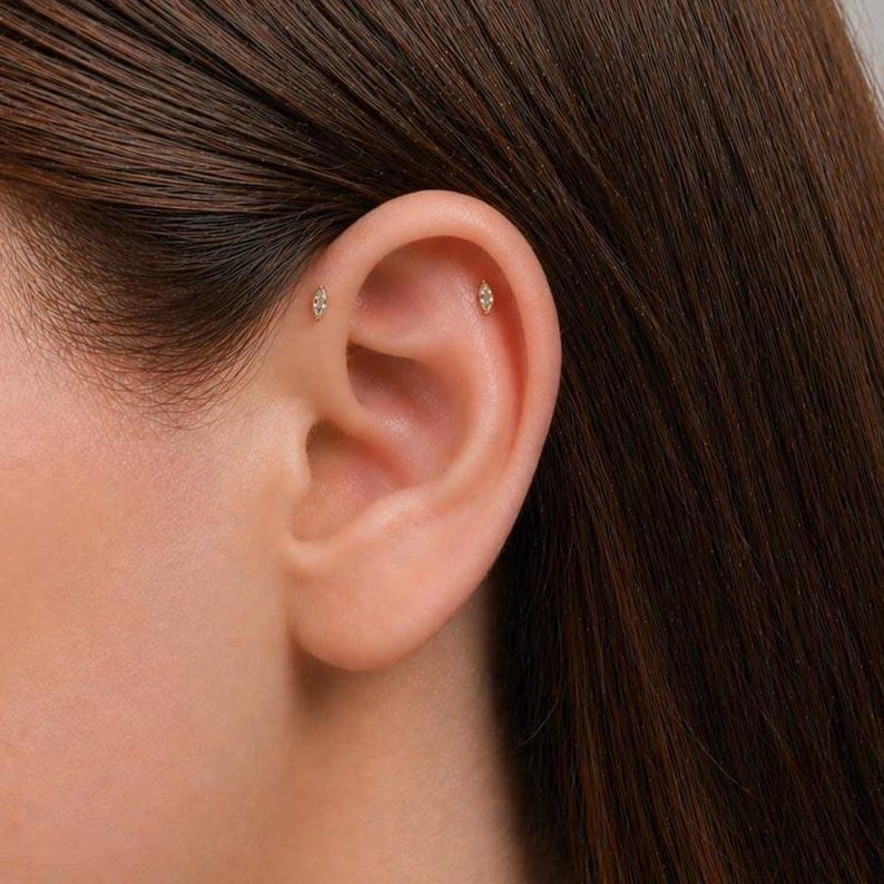 May include: Two small, marquise-shaped stud earrings made of a metallic material, each featuring a single clear stone. The studs are positioned in the upper cartilage area of the ear, with one placed slightly higher than the other.