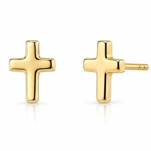 May include: Two gold-colored cross stud earrings. The earrings are displayed on a white background. The crosses have a simple, classic design with rounded edges. The earrings are likely made of a shiny metal, reflecting light.