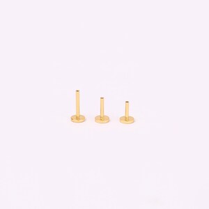May include: Three gold-colored flat back labret studs arranged in a row by height. Each piece features a circular base and a smooth, cylindrical post extending upwards. The metal has a polished, reflective finish, and the items are displayed upright.