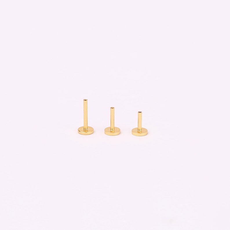 May include: Three gold-colored flat back labret studs arranged in a row by height. Each piece features a circular base and a smooth, cylindrical post extending upwards. The metal has a polished, reflective finish, and the items are displayed upright.