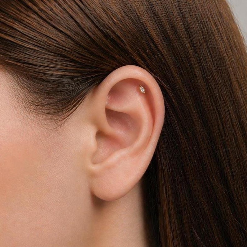 May include: A close-up of an ear with a small, oval stud earring. The earring is a light colour, possibly gold or silver, and has a small, clear stone. The ear is smooth and the skin is a light tone. The hair is brown.