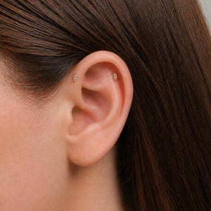 May include: Close-up of an ear with two small, gold-coloured earrings. Each earring has a diamond-shaped design with a central gemstone. The earrings are positioned on the upper ear cartilage.