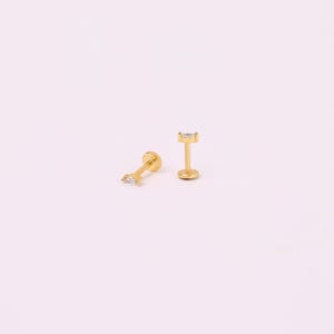 May include: Two gold-coloured, flat-back stud earrings, each with a rectangular, clear gemstone. One earring lies on its side, the other stands upright. The earrings are set against a light pink background.