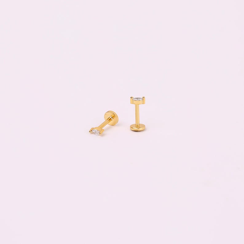 May include: Two gold-coloured, flat-back stud earrings, each with a rectangular, clear gemstone. One earring lies on its side, the other stands upright. The earrings are set against a light pink background.