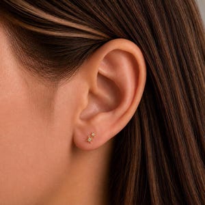 May include: A gold stud earring featuring two small, connected starburst shapes. The larger starburst is positioned slightly below the smaller one, and both are accented with tiny, clear, sparkling stones set in the center of each star point design.