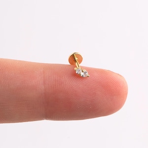 May include: A small, gold-colored earring with a flat back and a post. The earring features two clear, round gemstones set side-by-side. The earring is shown on a fingertip against a white background.