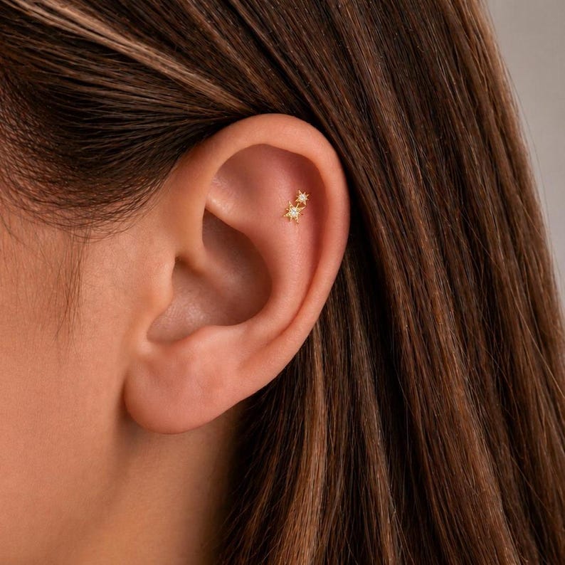 May include: A gold-toned ear stud featuring two connected starburst shapes, each accented with a small clear stone at its center. The jewelry is positioned in the upper cartilage area of the ear, showcasing a delicate and metallic design.