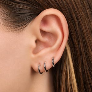 May include: Three small, polished silver-tone hoop earrings are worn in a row along the lower edge of the earlobe. Each earring is a simple, circular band with a smooth, reflective metallic finish, creating a clean and uniform appearance.