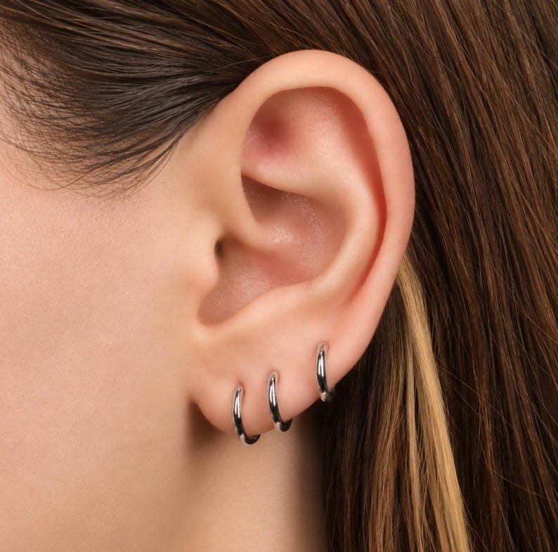 May include: Three small, polished silver-tone hoop earrings are worn in a row along the lower edge of the earlobe. Each earring is a simple, circular band with a smooth, reflective metallic finish, creating a clean and uniform appearance.
