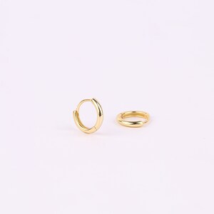 May include: A pair of small, polished gold hoop earrings. One earring is positioned upright in a circular shape, while the other lies flat on its side. Both hoops feature a smooth, reflective metallic surface and a simple hinged closure mechanism.