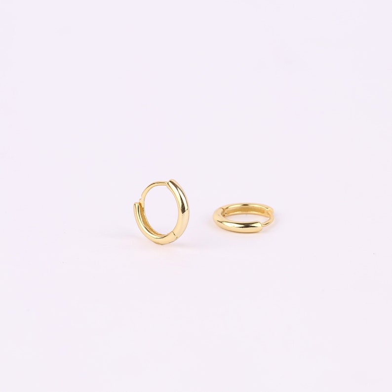May include: A pair of small, polished gold hoop earrings. One earring is positioned upright in a circular shape, while the other lies flat on its side. Both hoops feature a smooth, reflective metallic surface and a simple hinged closure mechanism.