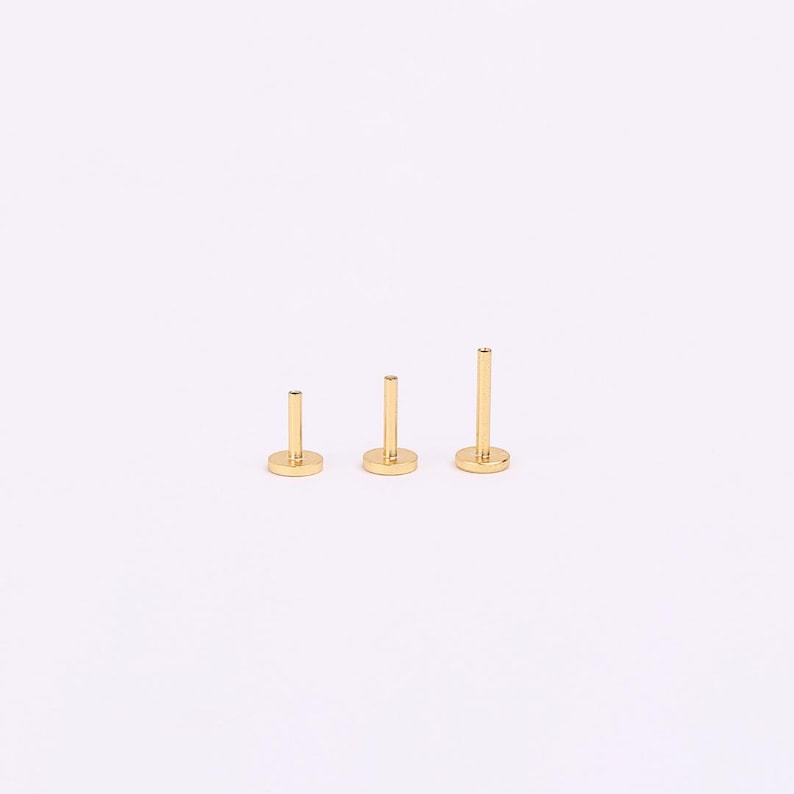 May include: Three gold-toned flat-back labret studs arranged in a row, each featuring a circular base and a vertical post of varying lengths. The studs are metallic with a smooth, polished finish, standing upright against a plain white background.