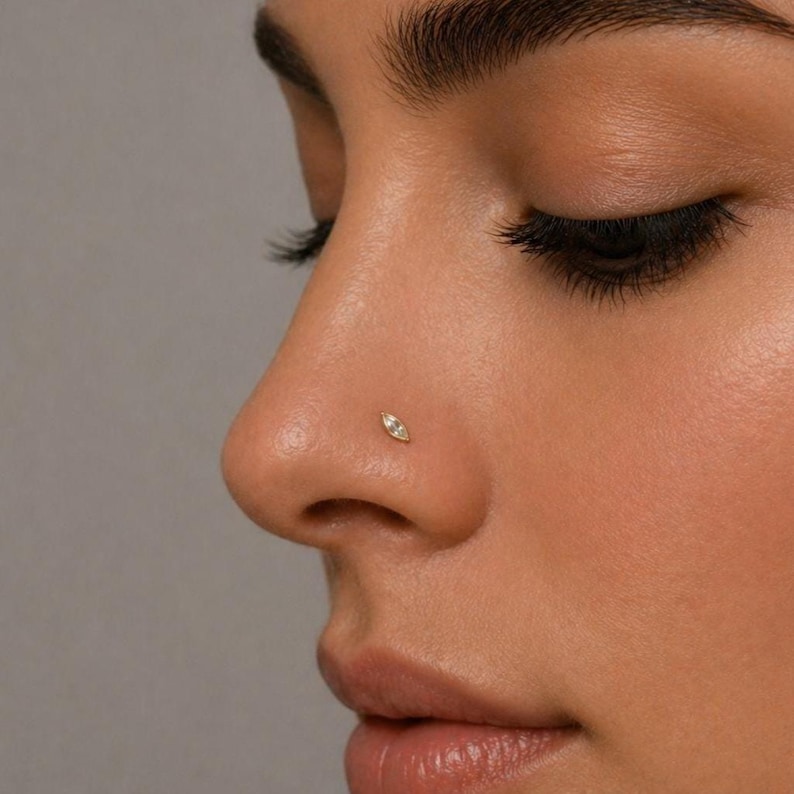 May include: A close-up of a nose with a small, gold-coloured nose stud. The stud is shaped like a marquise, with a small, clear stone in the centre. The skin tone is light, and the background is a solid grey.