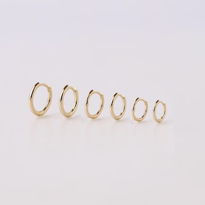 May include: A set of six gold hoop earrings arranged in a row from largest to smallest. Each earring features a polished, smooth, circular band with a hinged closure mechanism, showcasing a consistent metallic yellow finish across all pieces.