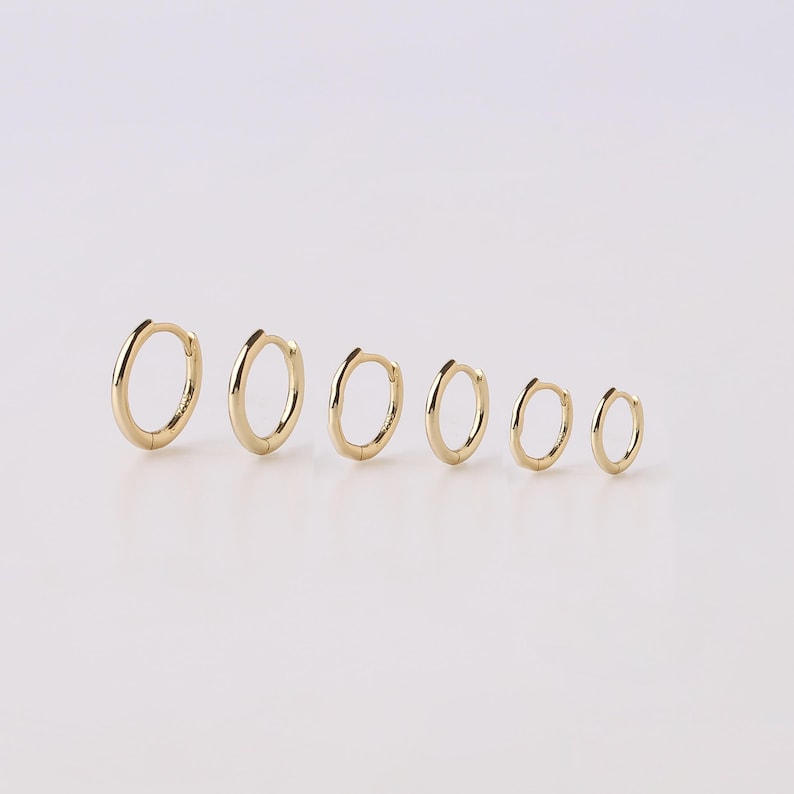 May include: A set of six gold hoop earrings arranged in a row from largest to smallest. Each earring features a polished, smooth, circular band with a hinged closure mechanism, showcasing a consistent metallic yellow finish across all pieces.