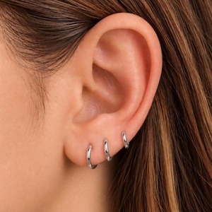 May include: Three small, polished silver-toned hoop earrings are worn in a row along the lower edge of the earlobe. Each earring features a simple, circular design with a smooth, reflective metallic finish, creating a uniform look.
