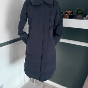 Puede incluir: Un abrigo largo azul marino con un cuello alto y redondeado. El abrigo tiene un diseño aerodinámico con un detalle de panel vertical en la parte delantera y bolsillos. El abrigo se muestra en un maniquí, mostrando toda su longitud y forma.