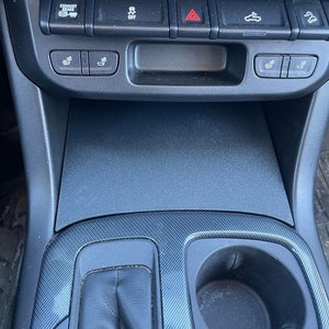 May include: A black car interior with a cup holder and a gear shift lever. The dashboard has several buttons, including an "Exhaust Brake" button, an "Off" button, a hazard light button, a button with a car icon, and a button with a person icon. There are also buttons for heated seats.